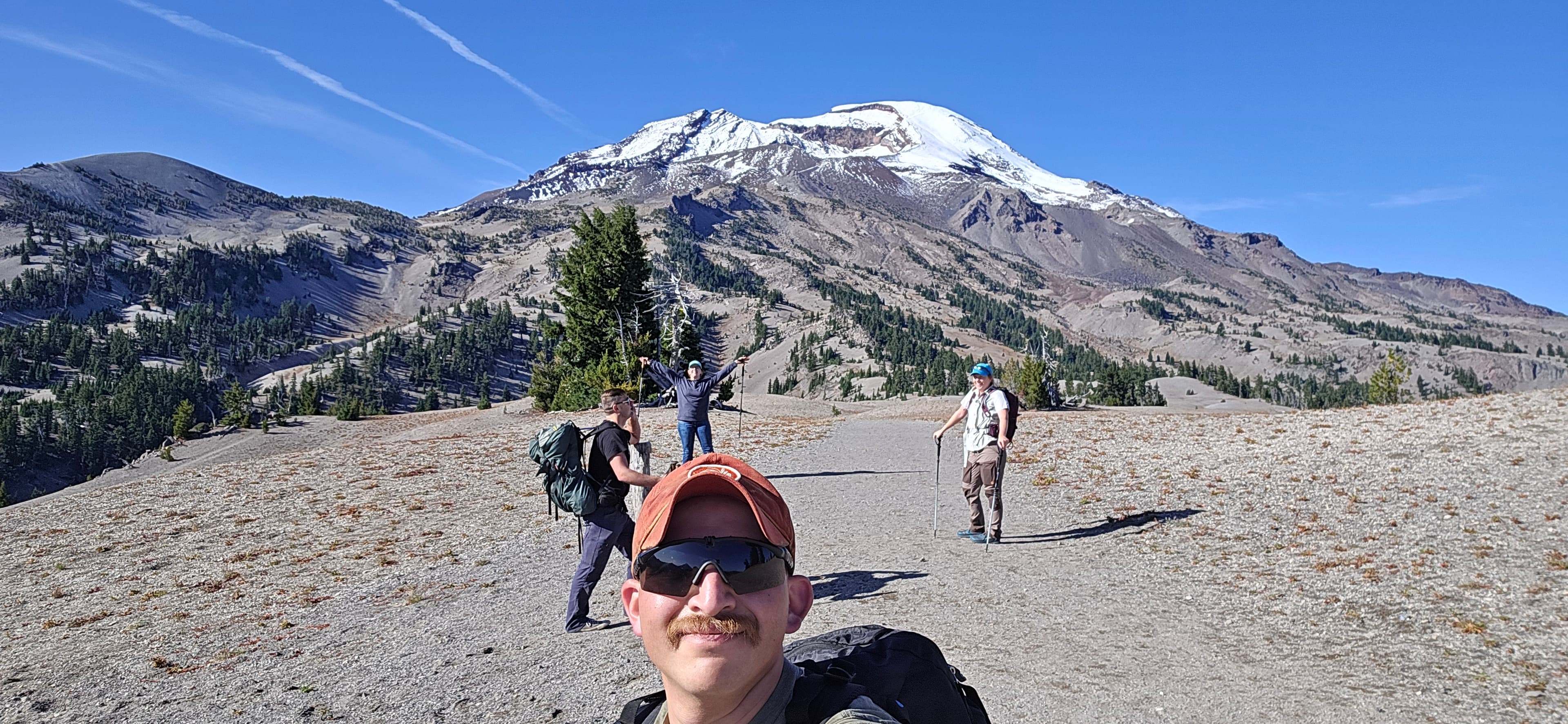 A hiker takes a summit selfie beneath a snowy ridge with two companions behind him.