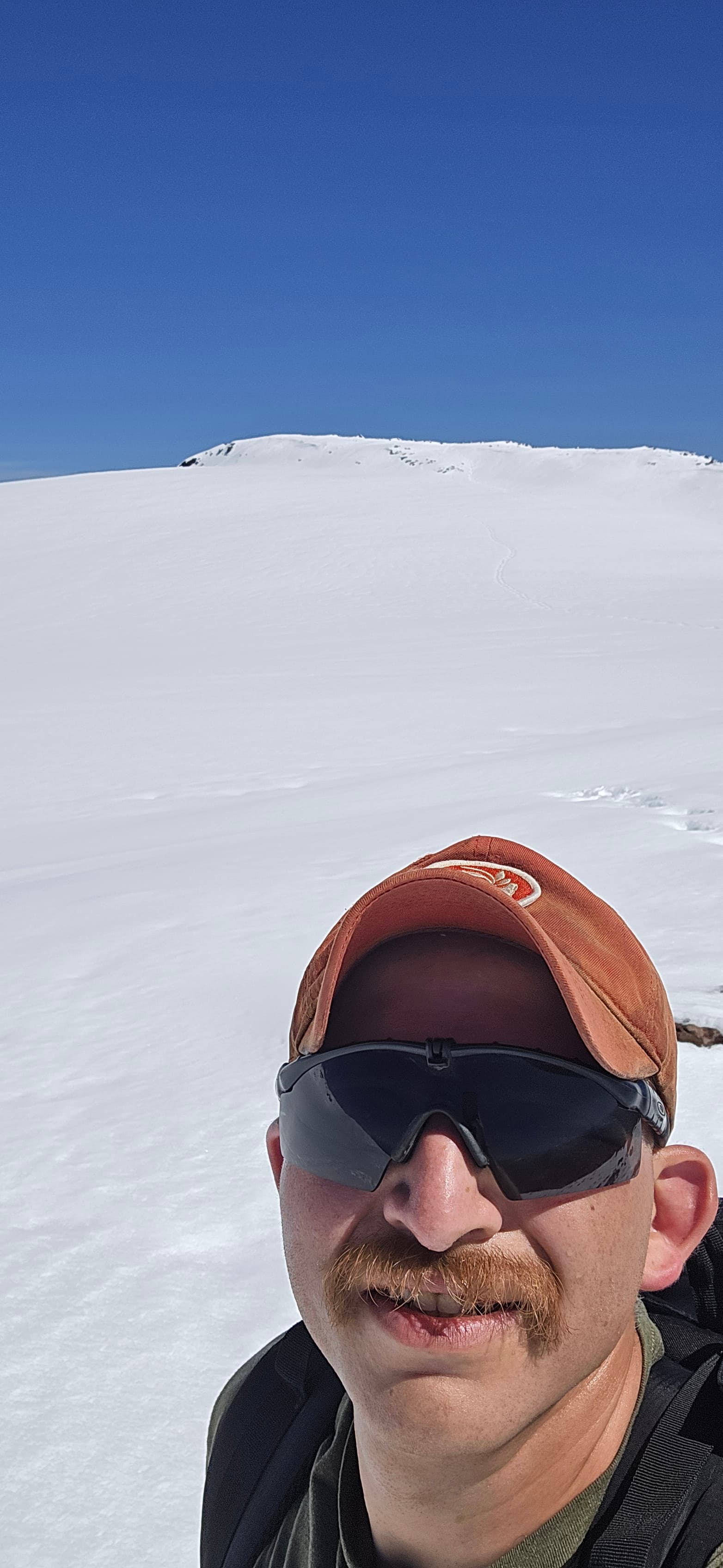 Close-up selfie in a red cap and sunglasses against a snowy mountain backdrop.
